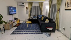 A modern living room with a black sofa, a small round table, a TV mounted on the wall, and a patterned rug. - Micasa-Sucasa Hotel and Suites (Port Harcourt)