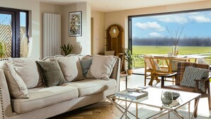 A living room with a sofa, coffee table, and a view of a green field through large windows. - Heron's Nest -  Ripple, Gloucester, Tewkesbury, sleeping two guests (Ripple)