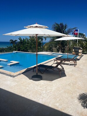 A swimming pool with lounge chairs and umbrellas, surrounded by palm trees and a view of the ocean. - Luxury BeachFront Villa with Private pool and Ocean View!
(Punta Rucia)