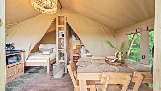 Una tienda de campaña con cama alta, una zona de comedor con mobiliario de madera y una lámpara colgante.