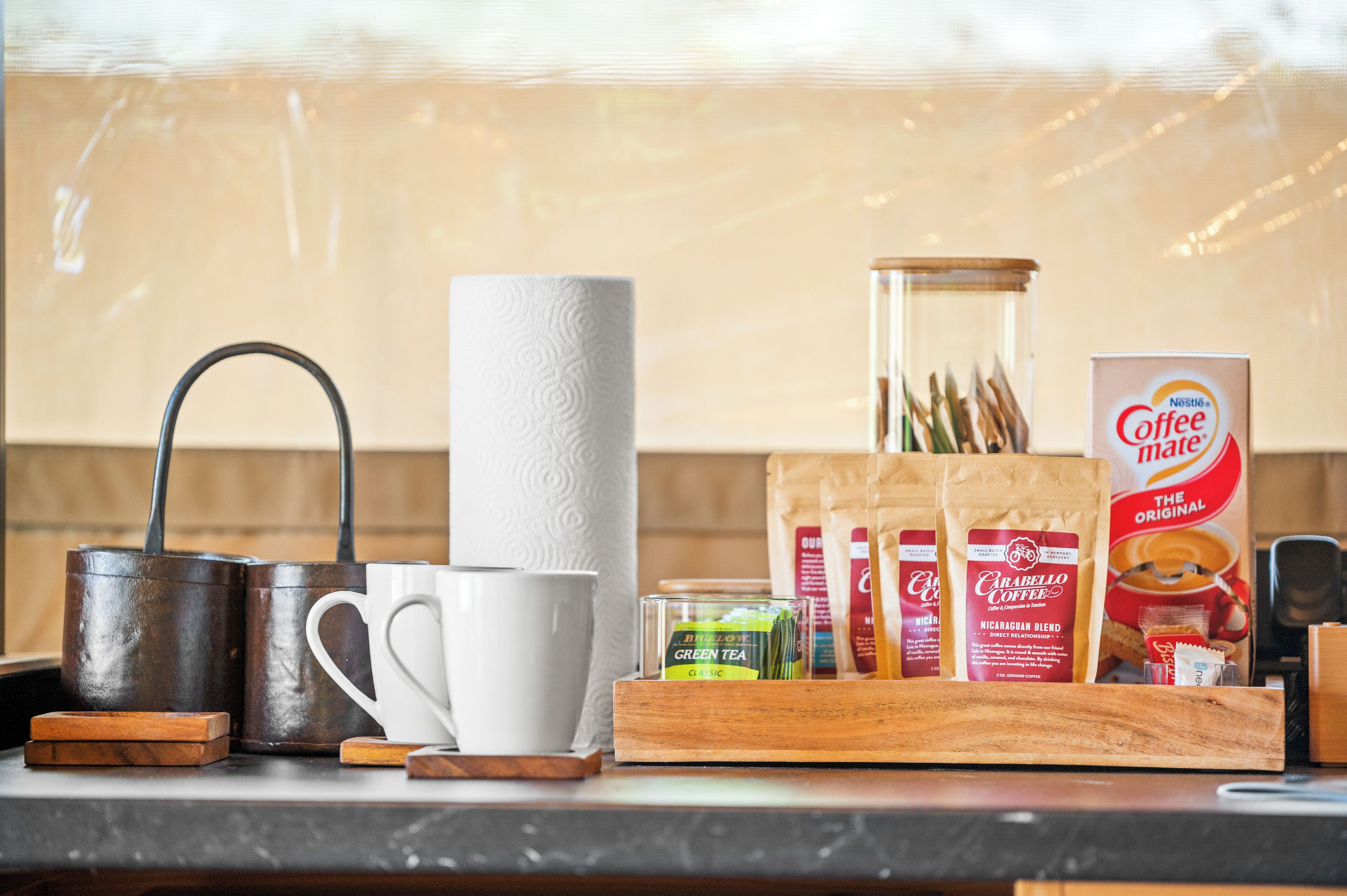 Una encimera de cocina con equipo para preparar café, una taza blanca, un rollo de papel de cocina, un tarro de cristal y paquetes de café.