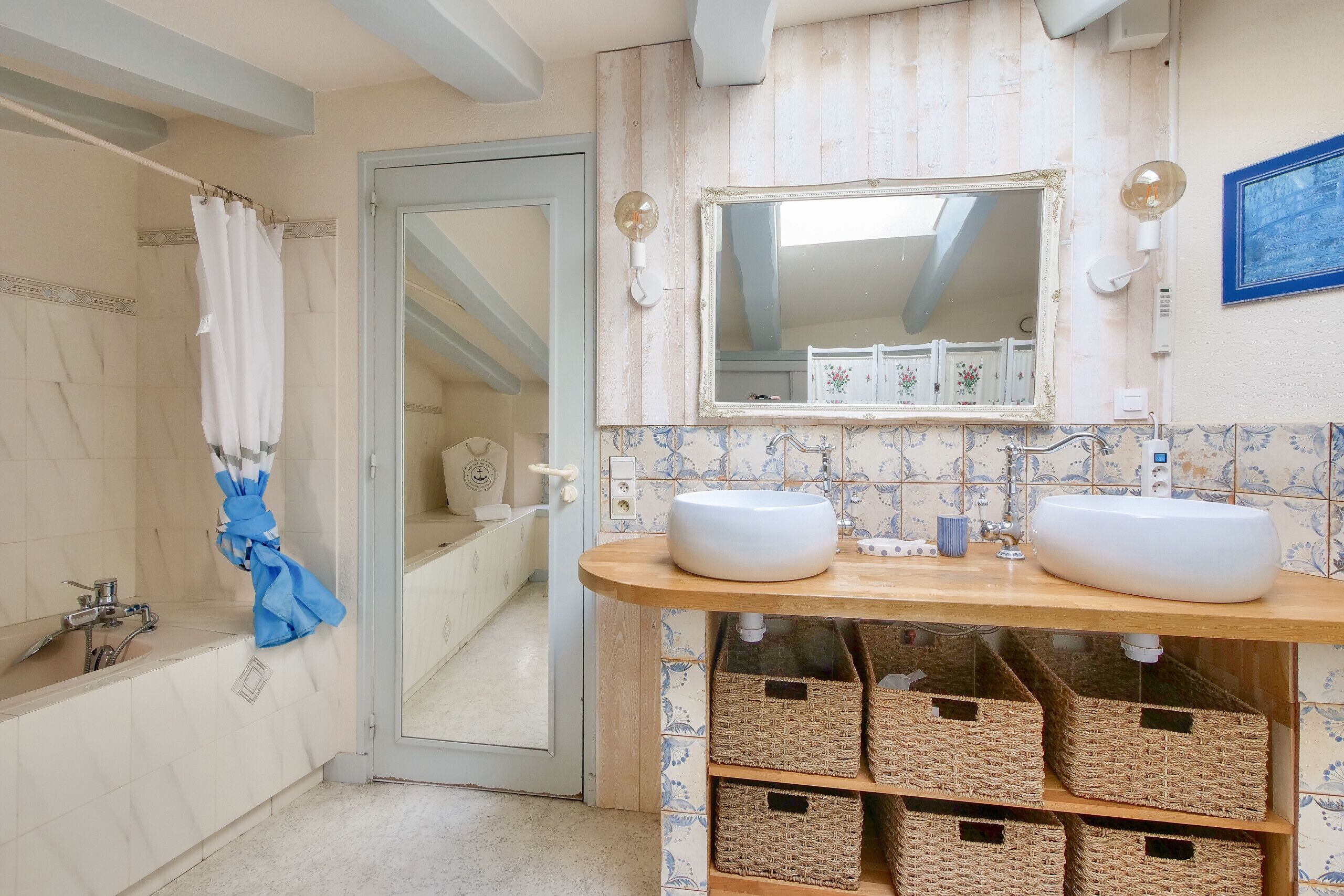 Une salle de bain avec deux lavabos, un miroir et une douche. On trouve des paniers en osier sous les lavabos et une serviette bleue est accrochée à la porte.