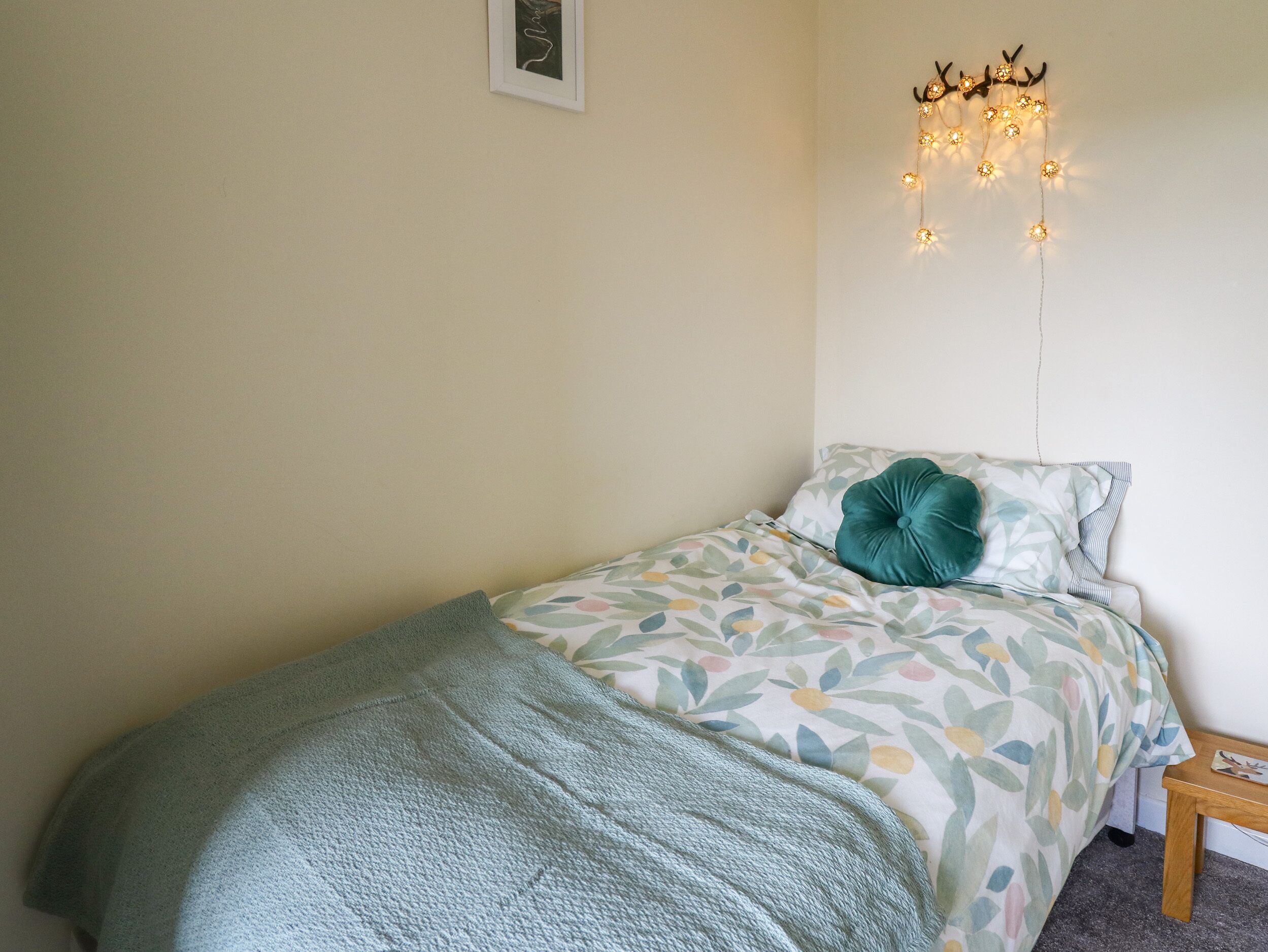 Een eenpersoonsbed met een bloemenprintdeken, een blauwgroen kussen en een lichtblauwe deken. Er staat een klein nachtkastje bij het bed en erboven hangt een ingelijst beeld aan de muur.