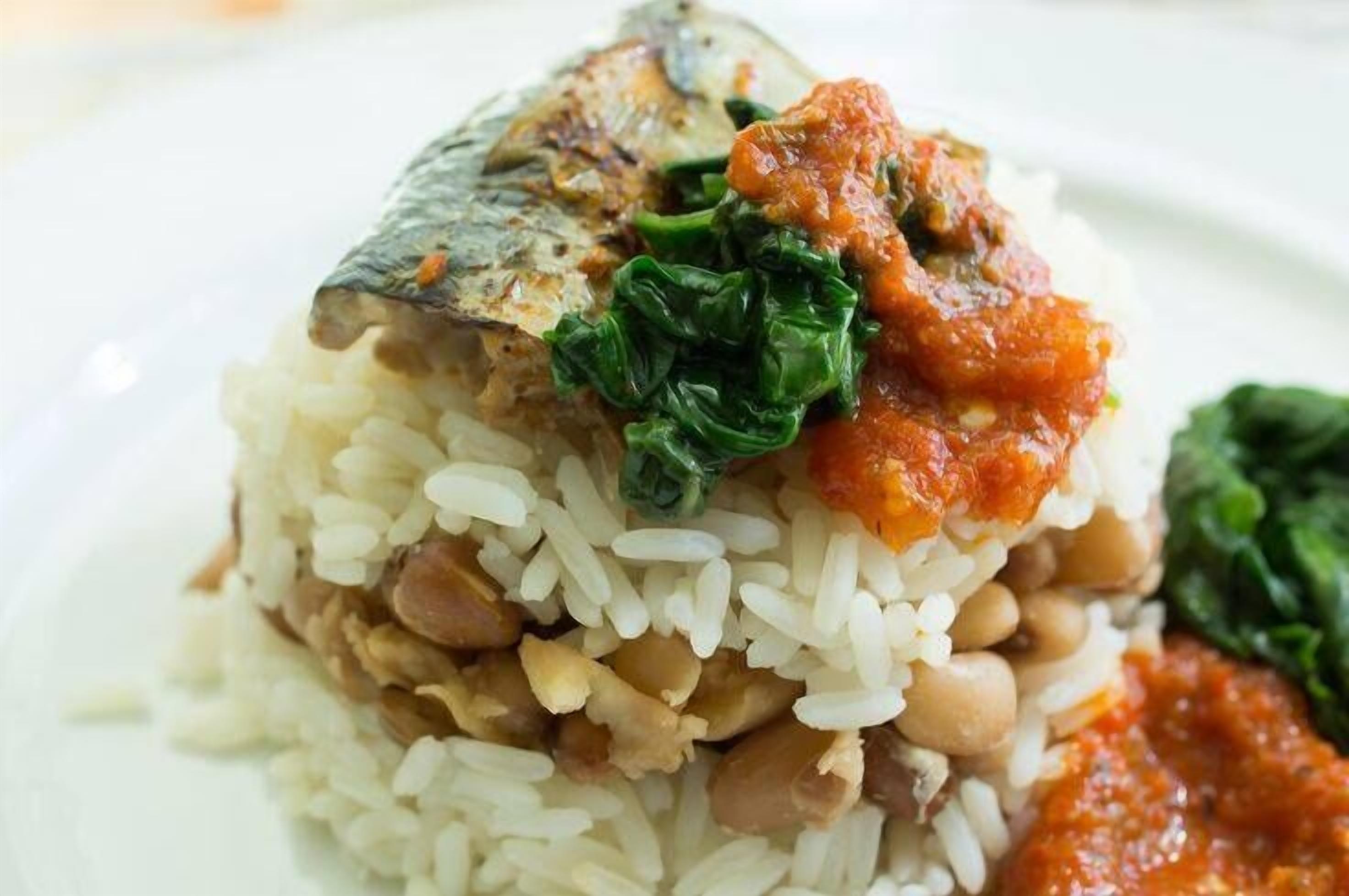 A plate with rice, beans, a piece of fish, and greens.