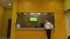 A reception area with a curved, ornate railing and a chandelier. A receptionist stands behind a counter with a television displaying a soccer game. - Limburg Hotel (Port Harcourt)