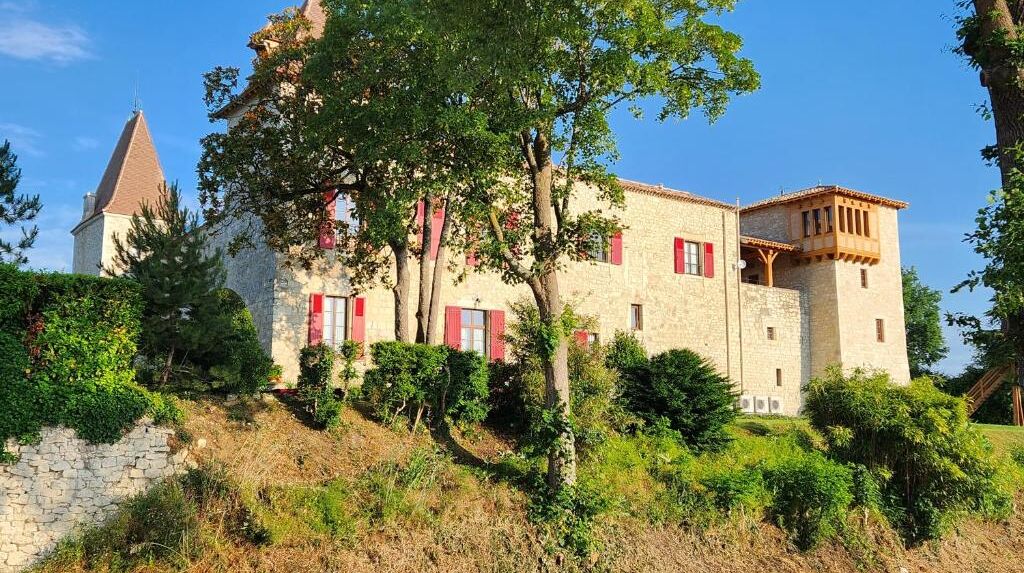 A stone building with red shutters, a tower, and a tree in front.
