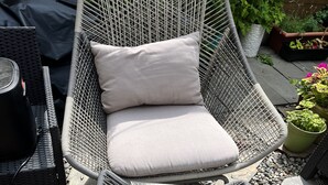 Une chaise d’extérieur en osier, dotée d’un coussin, placée sur une surface de gravier, avec des plantes en pot à proximité.