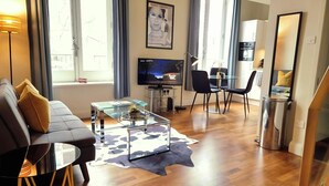 A modern living room with a sofa, coffee table, and TV. There is a dining area with chairs and a small table. The room has wooden flooring and large windows. - Notting Hill - The One One One! (3) (Greater London)