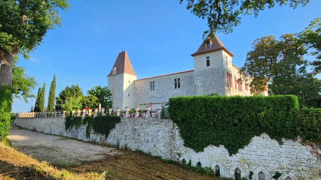 A historic stone building with two towers, a stone wall with greenery, and a clear blue sky.