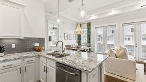 A modern kitchen with white cabinetry, granite countertops, and a built-in dishwasher. There is a dining area with a table and chairs, and a large window with blinds. - 30A:6-Seater Golf cart, Bikes, Beach Gear, 2 Pools (Inlet Beach)