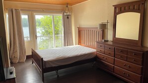A bedroom with a wooden bed, a dresser with drawers, a mirror, and a window with curtains. - Discover Melody Lodge, Ontario's Best-Kept Secret Where Tranquil Nature, Adventure, and Comfort Await. (Seeley's Bay)