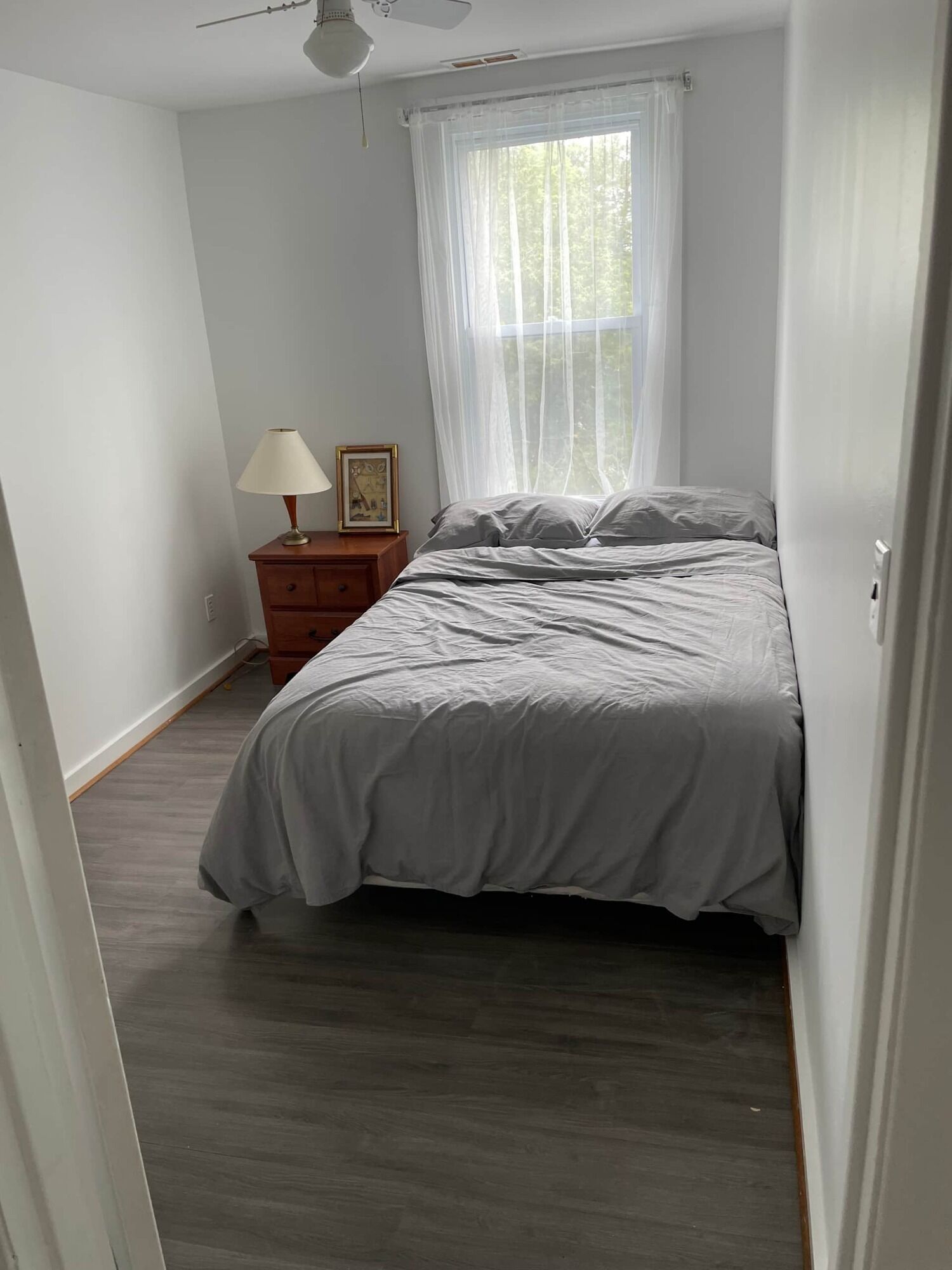 Een slaapkamer met een bed, een nachtkastje met een lamp, een lijst met een afbeelding en dunne gordijnen voor een raam.