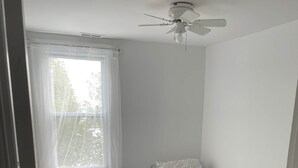 Ein kleiner, sauberer Raum mit zwei Einzelbetten, einem Deckenventilator und einem Fenster mit durchsichtigen Vorhängen.