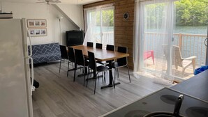 Una habitación con una mesa de comedor de madera, sillas negras, un ventilador de techo, un televisor y vistas a un lago a través de amplias ventanas.