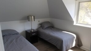 Un dormitorio con dos camas individuales, una mesita de noche con lámpara, y una ventana que deja entrar luz natural.