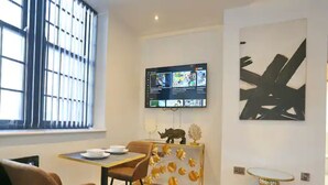 Ein zeitgemäßer Essbereich mit Tisch und Stühlen, einem an der Wand befestigten Flachbildfernseher und einem abstrakten Kunstwerk.