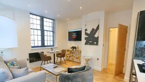 Ein modernes Wohnzimmer mit grauem Sofa, einem Couchtisch und einem grauen Sessel. Ein Fernseher ist an der Wand befestigt, ein Essbereich mit Stühlen vorhanden, und ein großes Fenster mit Jalousien.