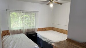 A compact bedroom with two single beds, a wooden dresser, a ceiling fan, and a window with sheer curtains. - Discover Melody Lodge, Ontario's Best-Kept Secret Where Tranquil Nature, Adventure, and Comfort Await. (Seeley's Bay)