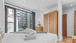Ein modernes Schlafzimmer mit einem großen Bett, einem Holzschrank und Blick auf einen Hochhaus durch eine Glastür.