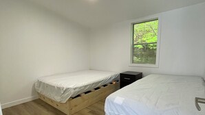 Et lille, rent værelse med to træsenge, et lille natbord og et vindue med udsigt til grønne områder.