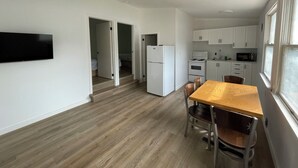 Et lille køkken med hvide skabe, en mikrobølgeovn og en komfur. Der er et spisebord med stole og et TV ophængt på væggen.
