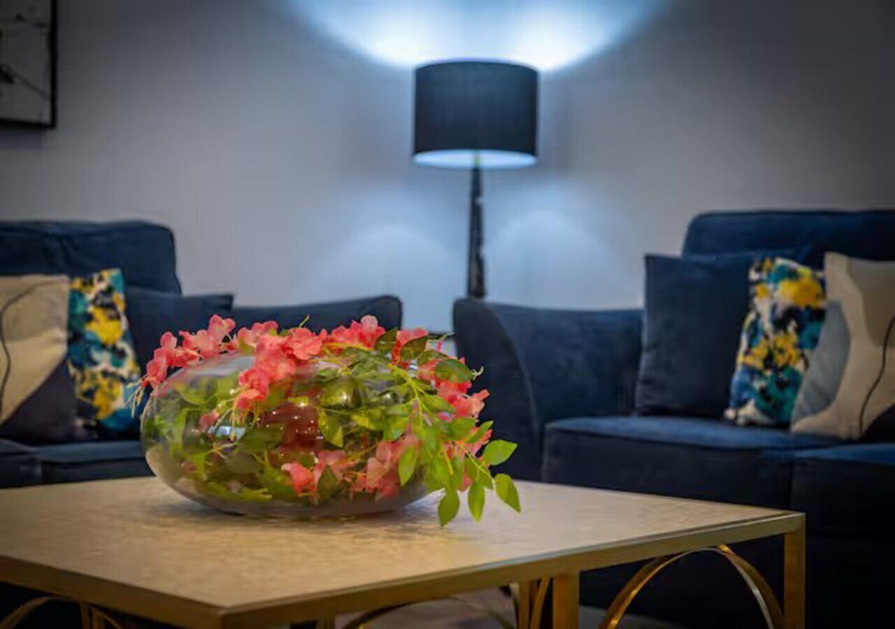 Un salon avec un vase en verre rempli de fleurs roses posé sur une table basse en bois, des canapés bleu foncé ornés de coussins colorés et un lampadaire noir.