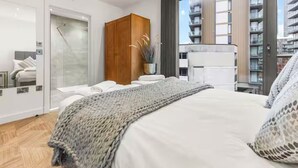 Ein modernes Schlafzimmer mit einem großen Bett, einem Holzschrank und Blick auf eine Stadtlandschaft durch ein großes Fenster.