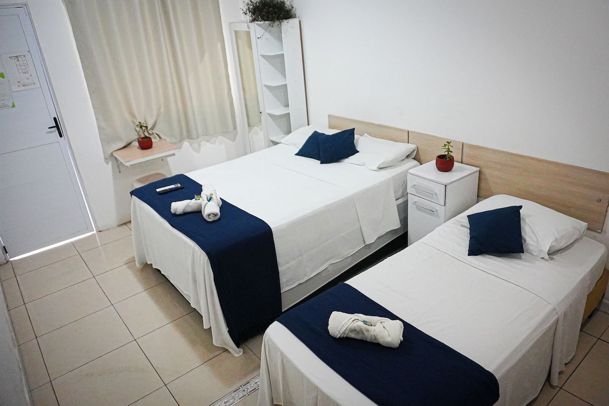 A hotel room with two beds, each with a blue bedspread and white linens, a bedside table with a red flower pot, and a white wardrobe.