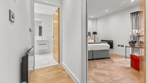 Una habitación de hotel moderna con suelo de madera, una cama con cabecero gris, una mesita de noche con lámpara y un taburete pequeño rojo.