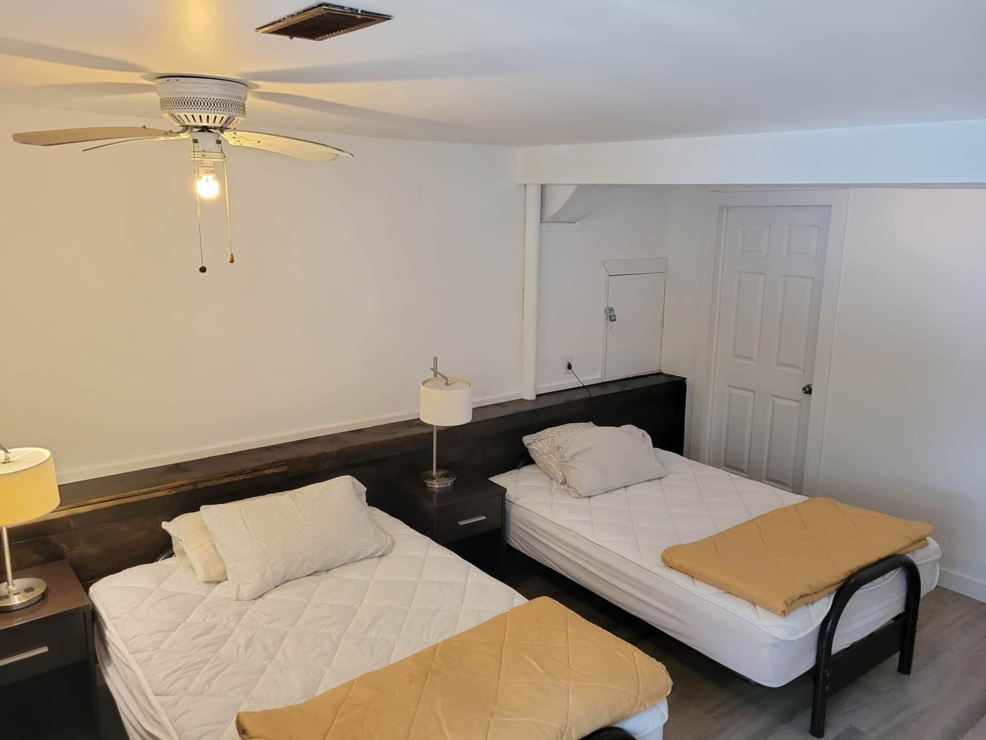 A bedroom with two single beds, a ceiling fan, a bedside table with a lamp, and a wardrobe.