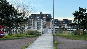 A wooden boardwalk leading to a gated entrance with modern apartment buildings in the background. - Appartement Cosy vue Port (Dives-sur-Mer)