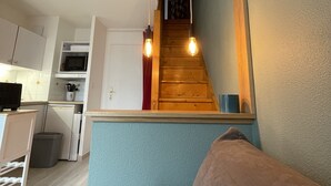 A compact living space with a kitchenette, a wooden staircase, and a seating area with cushions. - Appartement Cosy vue Port (Dives-sur-Mer)