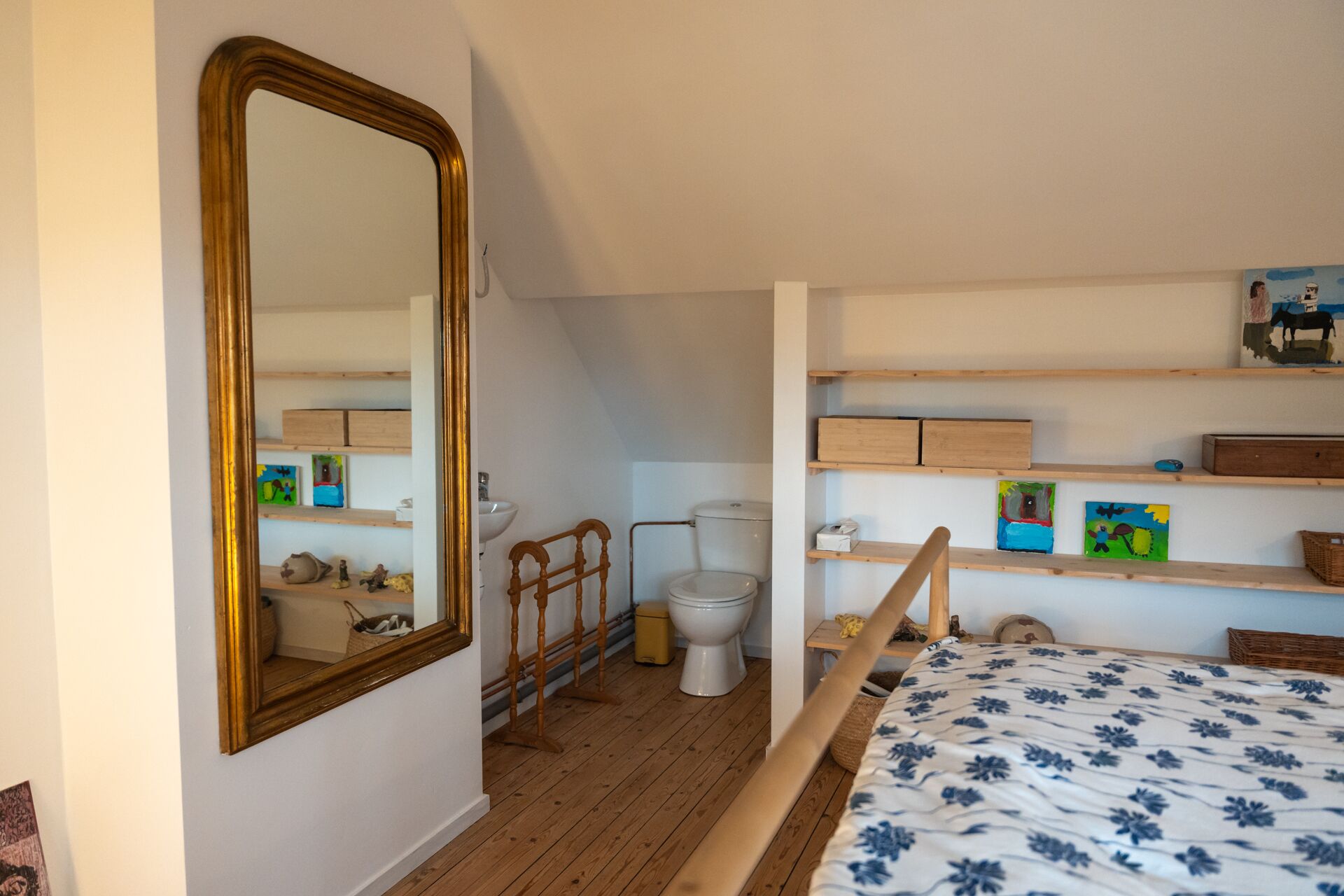 Una camera da letto con pavimento in legno, un letto con copripiumino floreale, uno specchio grande, un water e mensole con diversi oggetti.