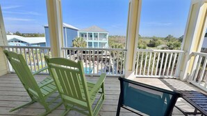 A wooden deck with two green chairs and a blue table, overlooking a pool and a light blue house. - A Beach Life (Gulf Shores)
