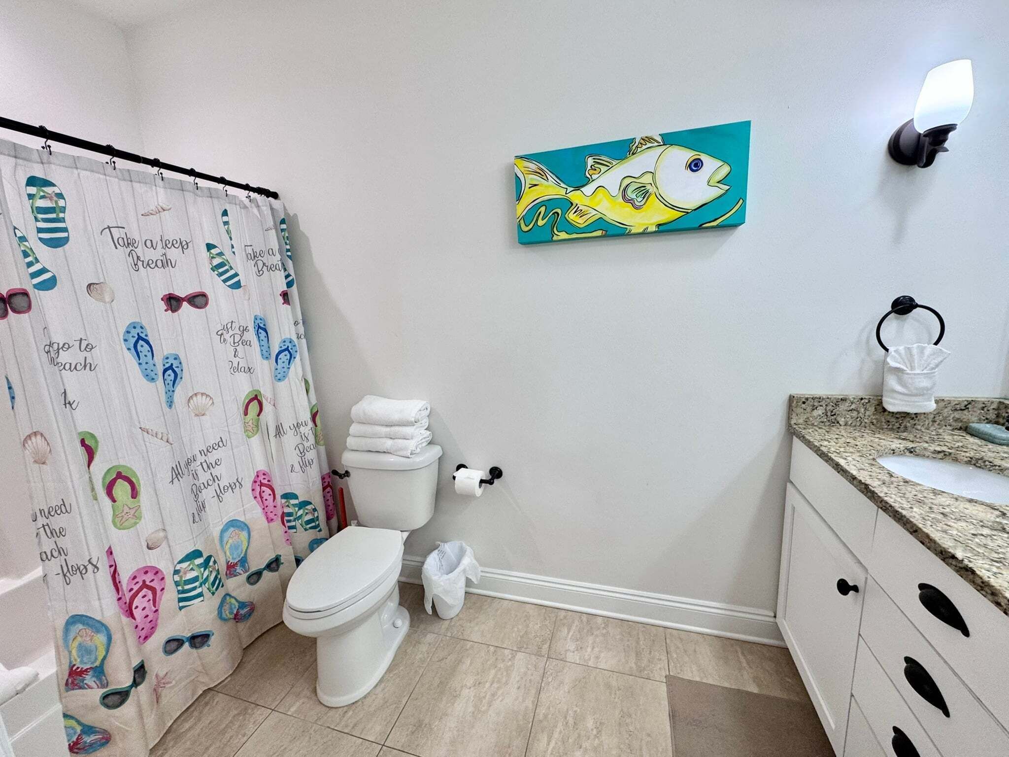 A bathroom with a shower curtain featuring beach and flip-flop designs, a toilet, a sink with a towel, and a wall-mounted light fixture.