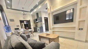 A modern living room with a grey sofa, a wooden coffee table, a TV mounted on the wall, and a kitchen area in the background. - VillaMira GuestHouse 2 Downtown Amman (Amman)