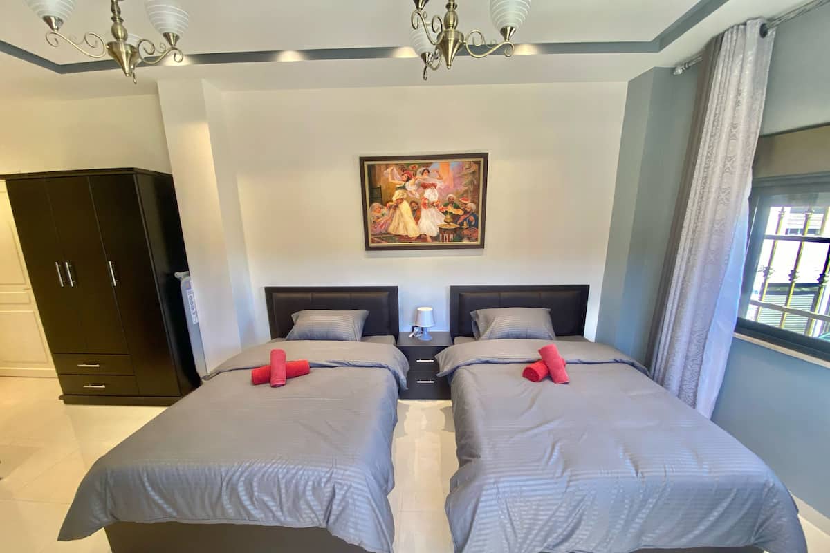 A double bed with grey linens and red towels, a painting above the bed, a wardrobe, and a window with curtains.