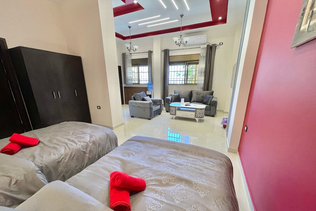 A hotel room with two beds, a dark wardrobe, a red wall, and a view of the living area with a sofa and a coffee table.