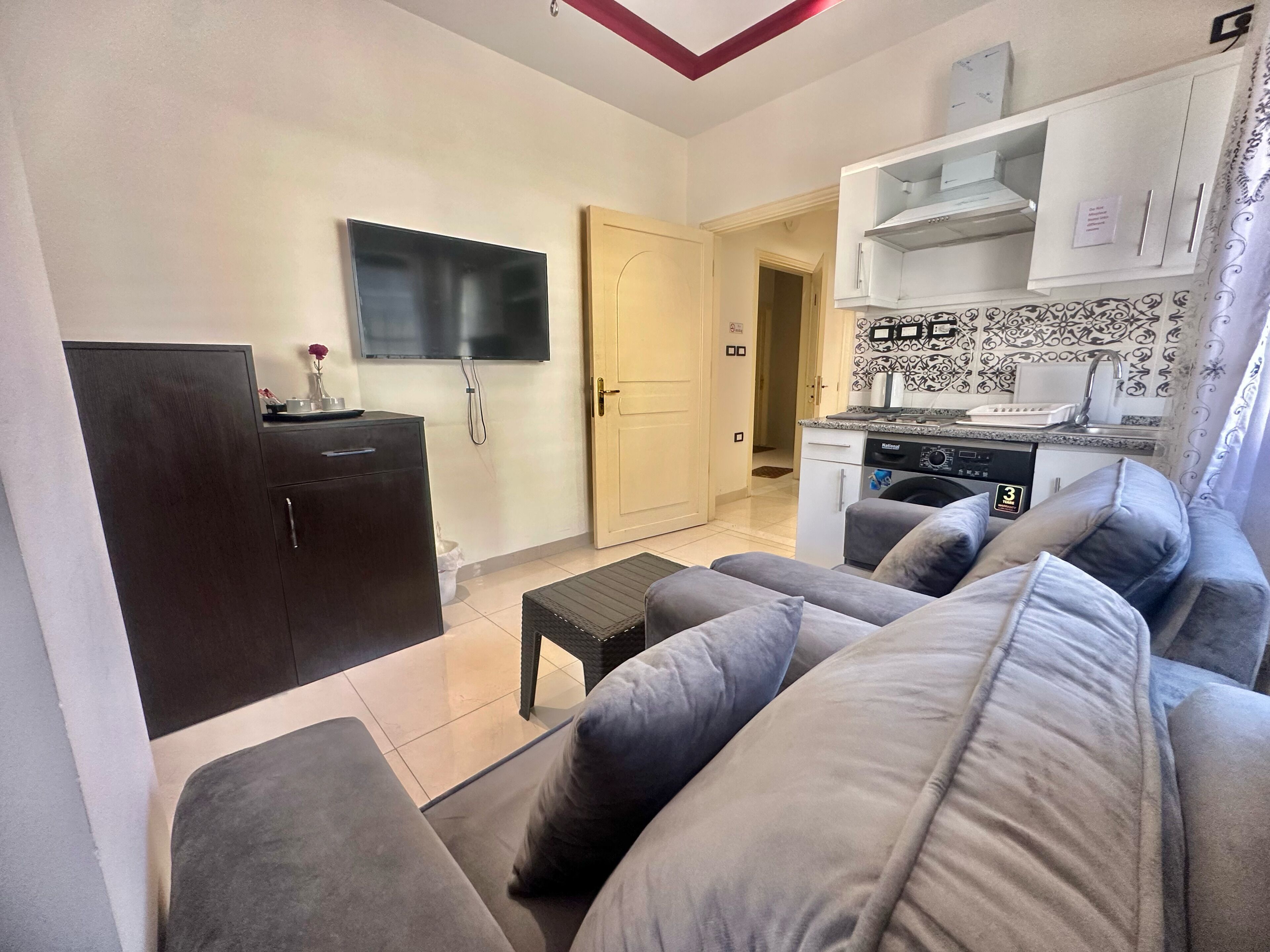 A compact living space with a sofa, a flat-screen TV, a dark wooden cabinet, a washing machine, and a kitchenette.