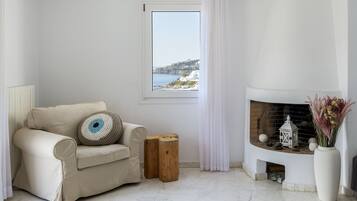 A modern living room with a beige sofa, a small wooden stool, a white fireplace, and a view of the sea through a window.