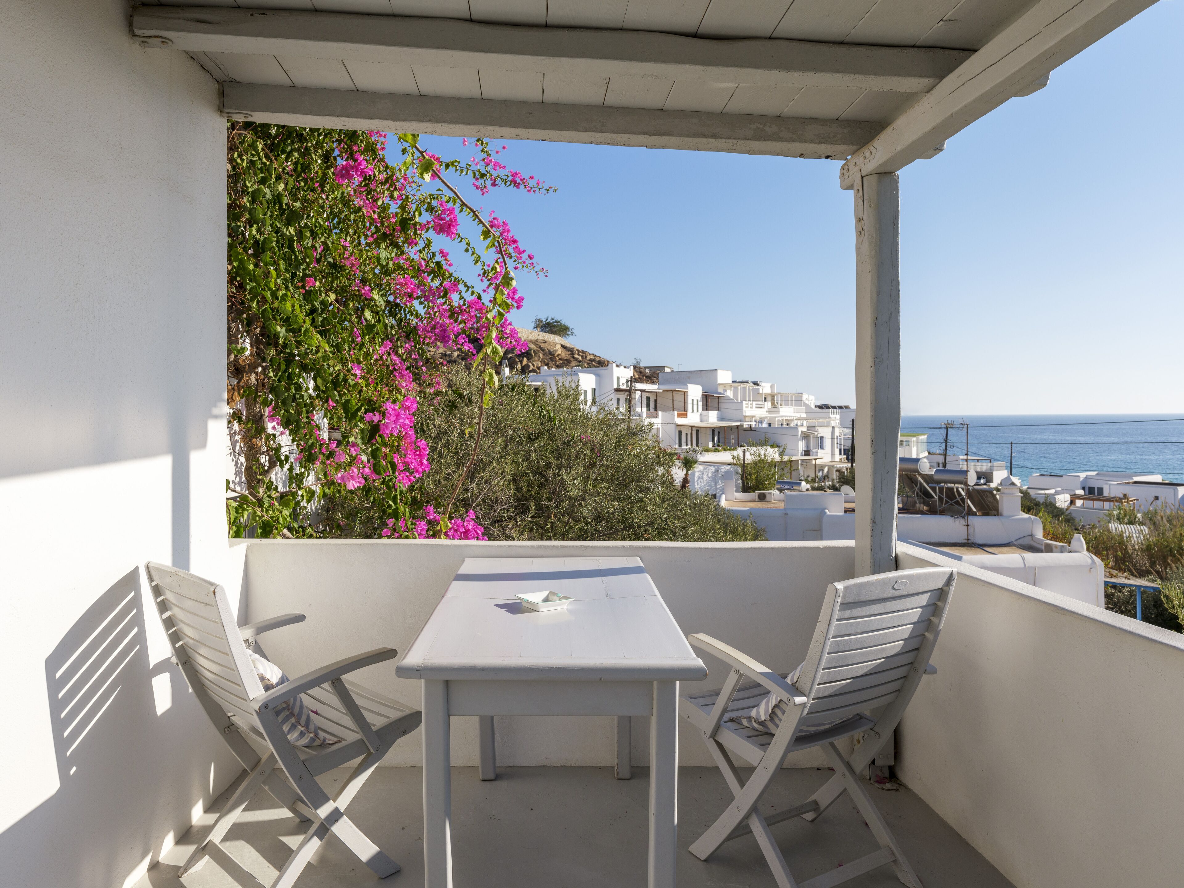 A balcony with a white table and two chairs, overlooking a coastal town with white buildings and the sea.