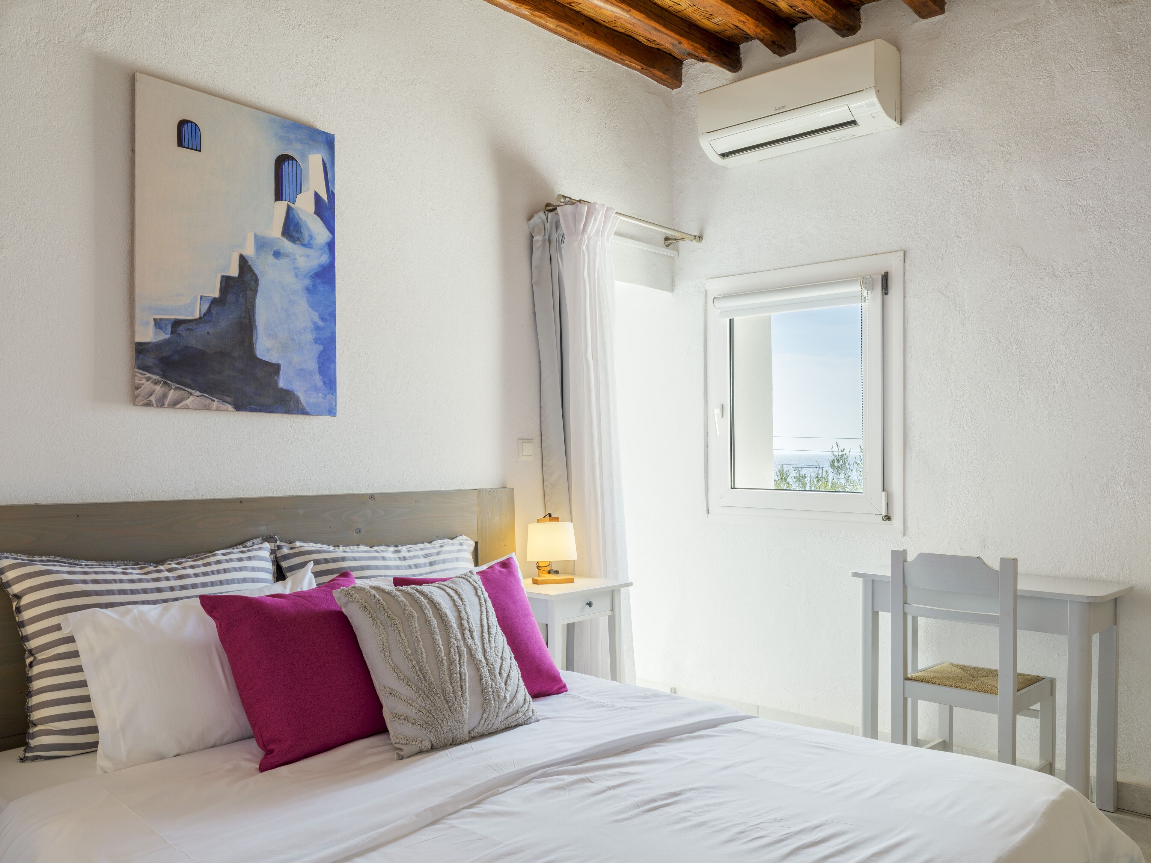 Un dormitorio con cama, almohadas, mesita de noche, lámpara, ventana y un cuadro en la pared.