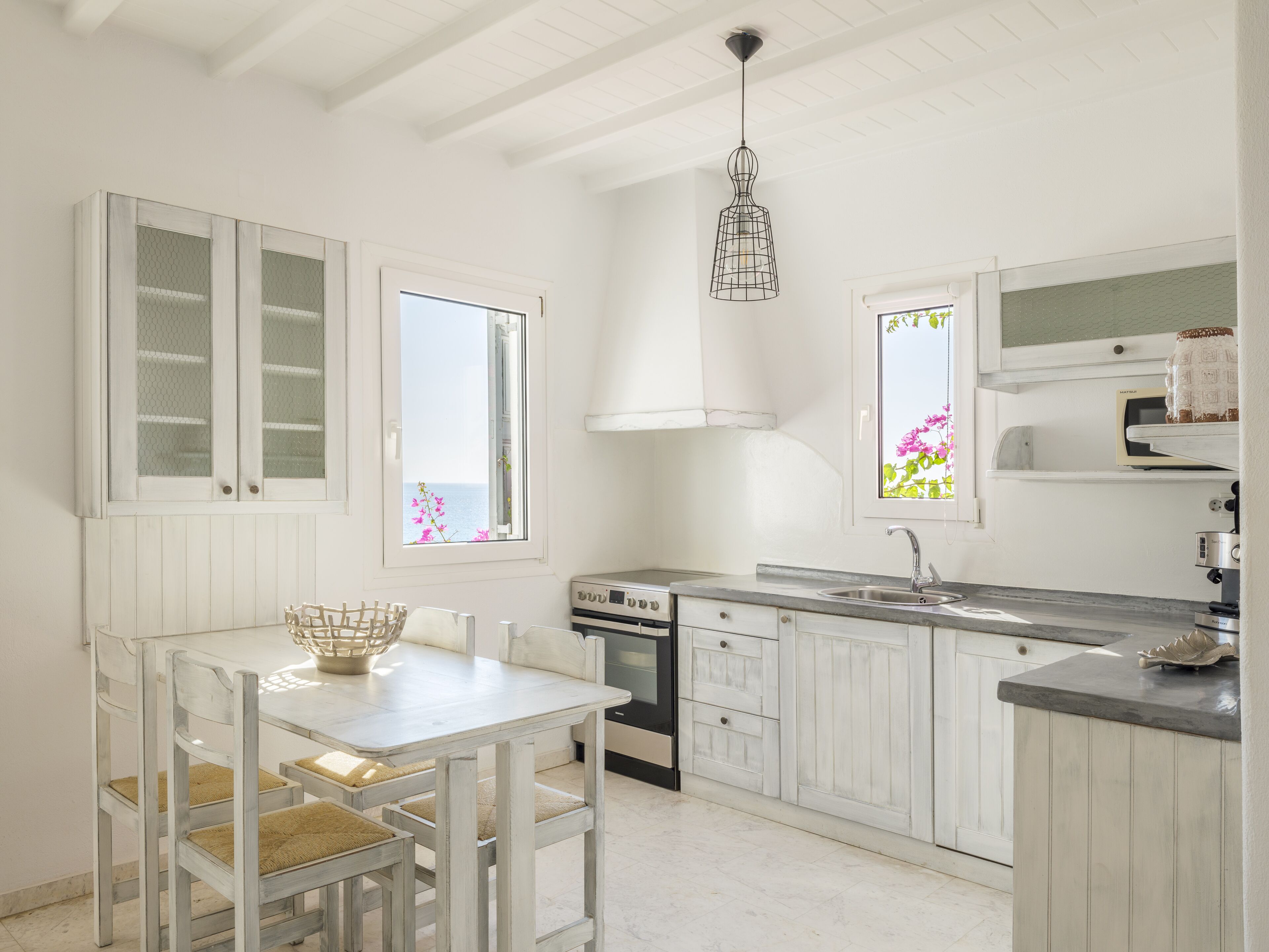 A kitchen with white cabinetry, a dining table with chairs, a hanging light fixture, and a window with a view of the sea.