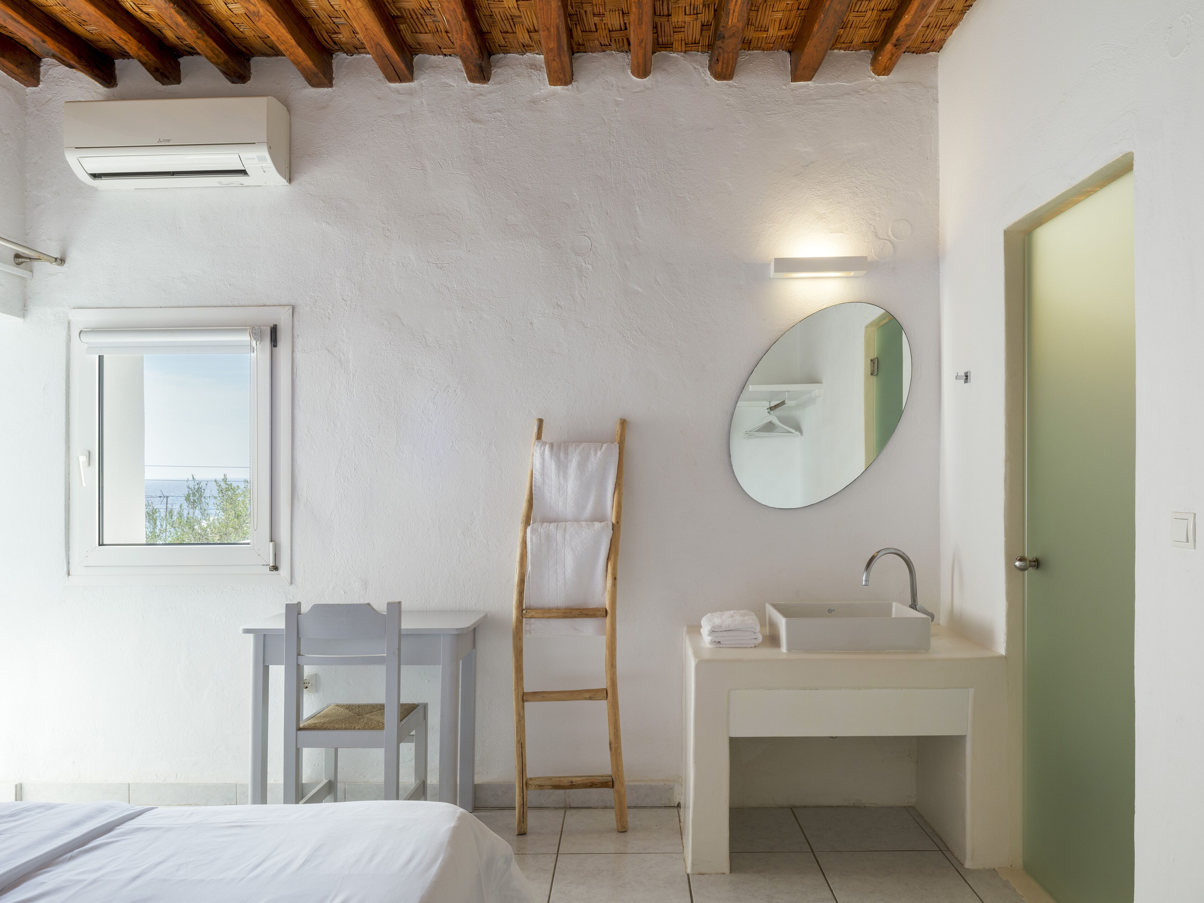 Un dormitorio con cama, ventana con vistas, escalera de madera, mesita de noche y baño con lavamanos y espejo.
