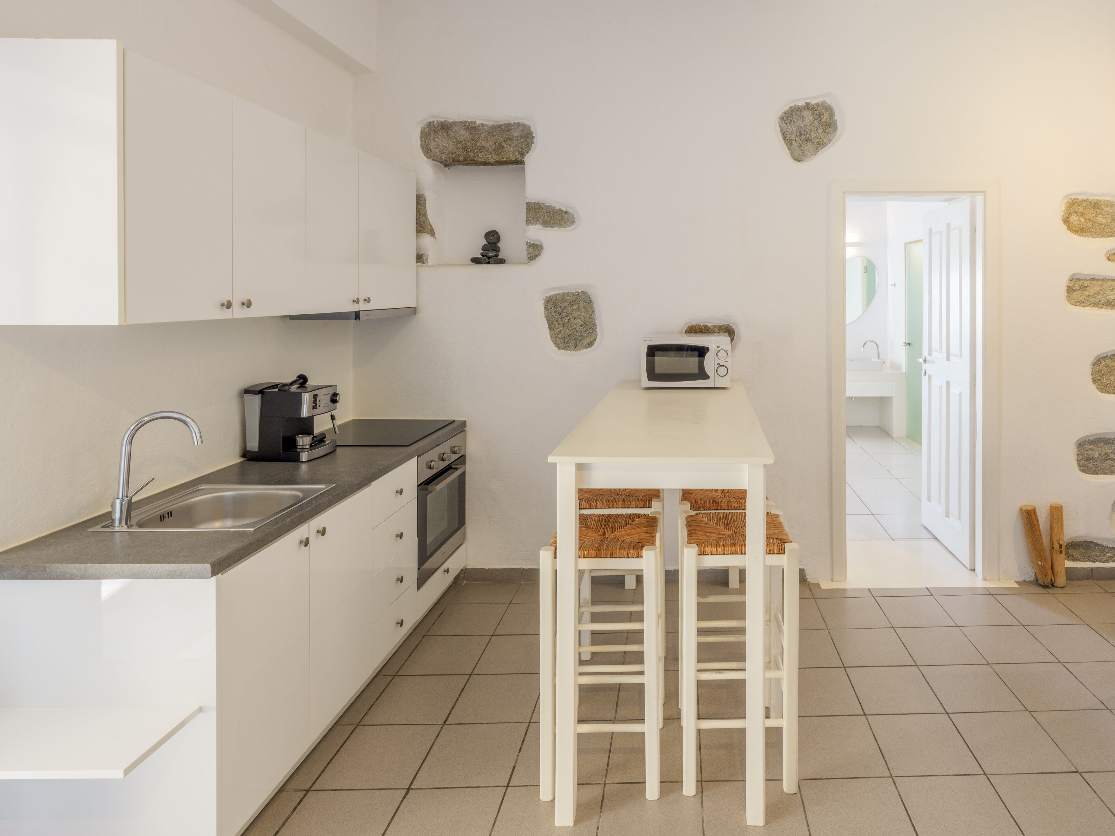 A modern kitchen with white cabinets, a stainless steel sink, a built-in oven, a microwave, and a small island with stools.