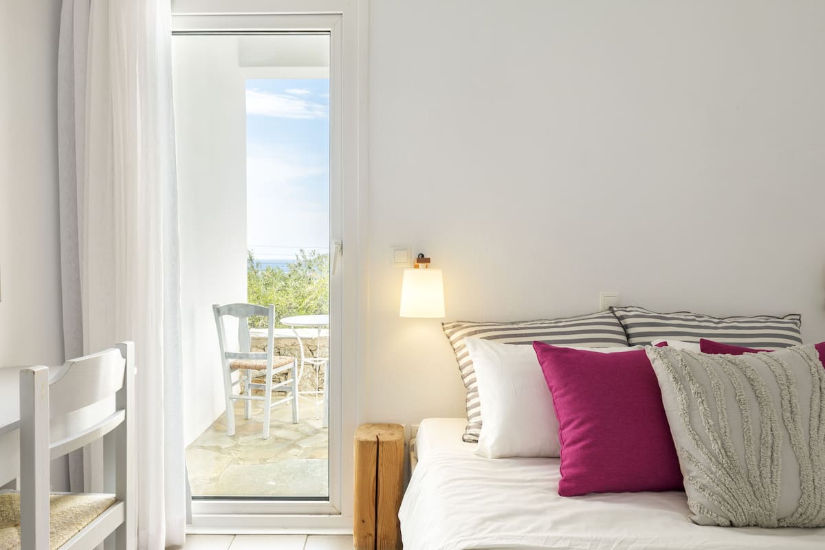 A bedroom with a bed, pillows, a bedside lamp, a chair, and a view of an outdoor patio.