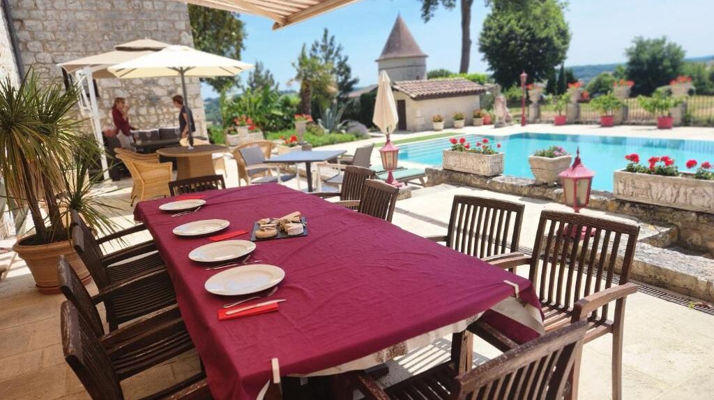Un área de comedor al aire libre con una mesa preparada para cuatro personas, rodeada de sillas de madera, cerca de una piscina.