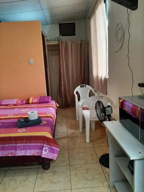 Une petite pièce, de taille modeste, comprenant un lit, un bureau, une chaise et un ventilateur.