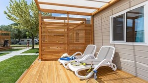 A covered outdoor area with wooden flooring, two white sun loungers, and a tennis racket leaning against one. - Vacation resort Oliva Mobilehome Premium for 4 persons (Rabac)