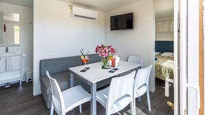 A compact room with a white table, two white chairs, a grey sofa, a flat-screen TV mounted on the wall, and a small kitchenette area with a sink and cabinets. - Vacation resort Oliva Mobilehome Premium for 4 persons (Rabac)